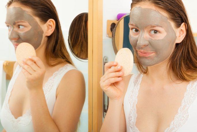 Beauty procedures spa and skin care concept. Young woman with facial clay mask in bathroom holds sponge to remove mud